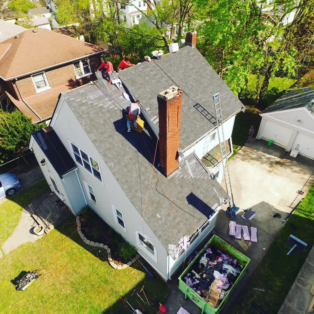 Roofers on a gray shingle roof beside a brick chimney, seen from overhead