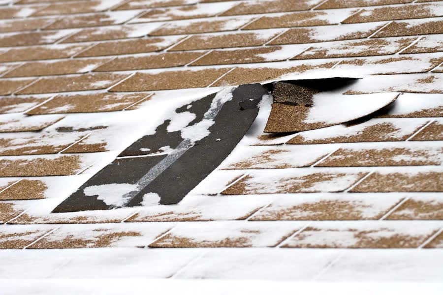 A torn shingle lifting from a roof dusted with snow